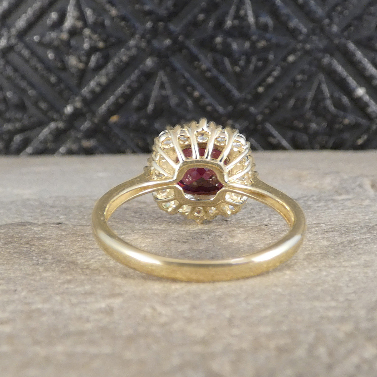 A yellow gold ring featuring a central cushion garnet gemstone surrounded by a halo of small clear stones, resting on a textured stone surface with a dark patterned background.