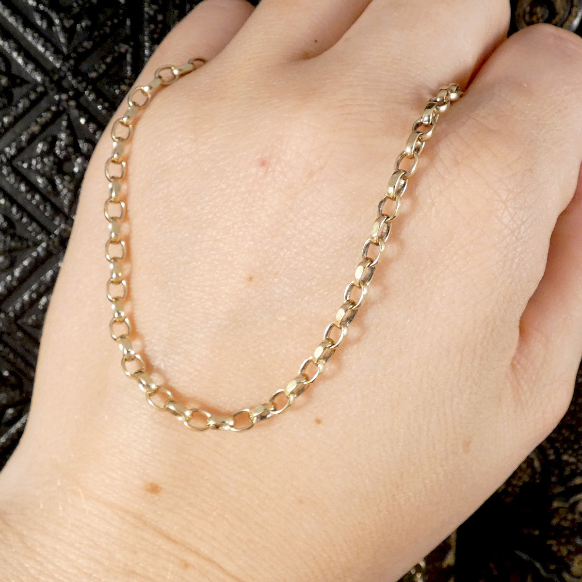 A close-up of a gold chain necklace draped across a person's hand, showing the chain's link pattern against a dark textured background.