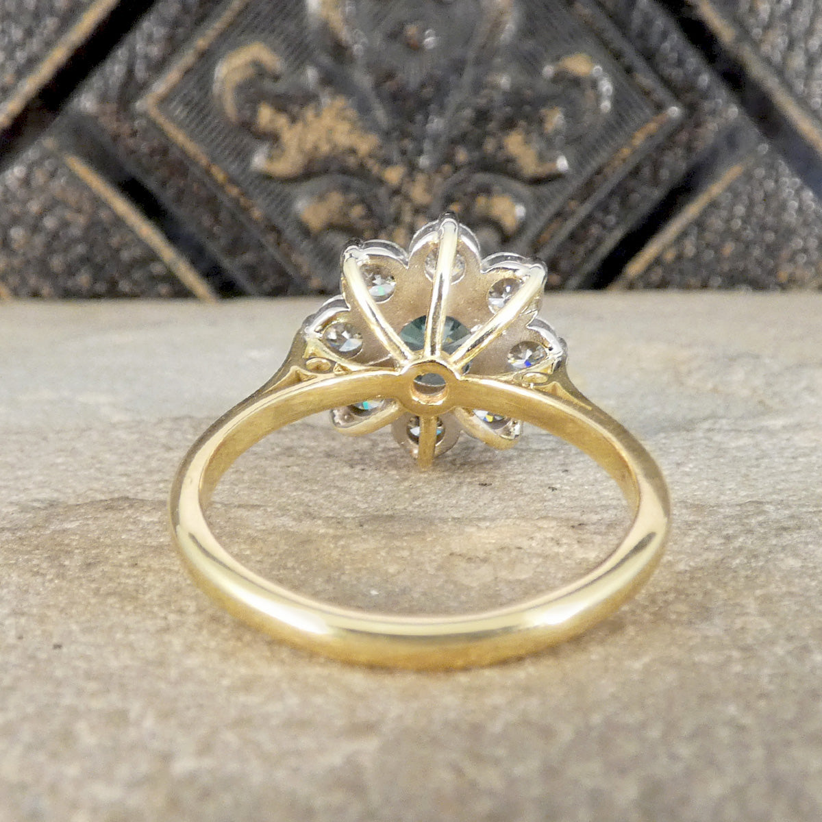 A yellow gold ring with a floral or sunburst cluster of small clear stones, photographed close-up on a stone surface with a dark decorative background.