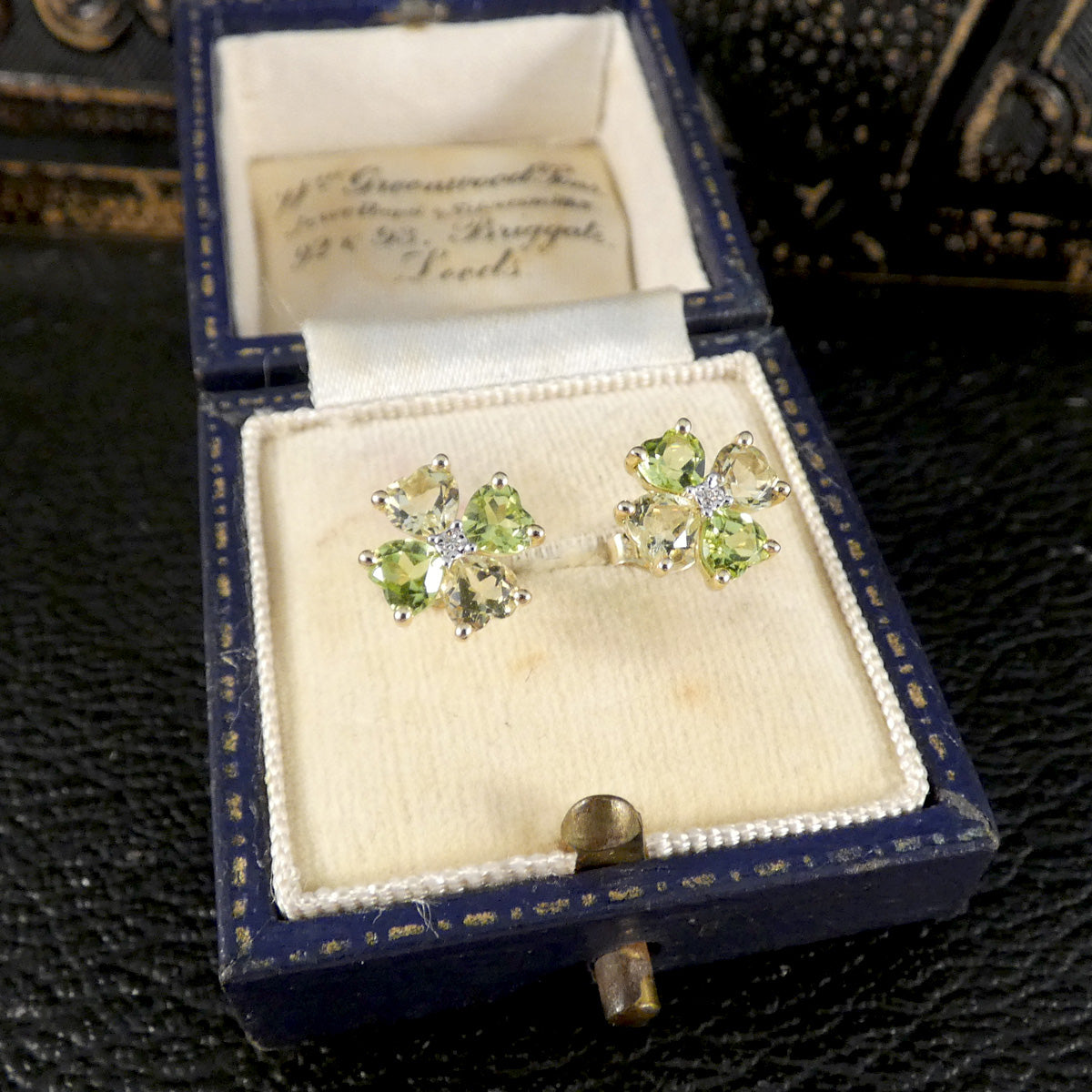 A pair of yellow gold stud earrings with four green and yellow gemstones arranged in a flower shape, in jewellery box.