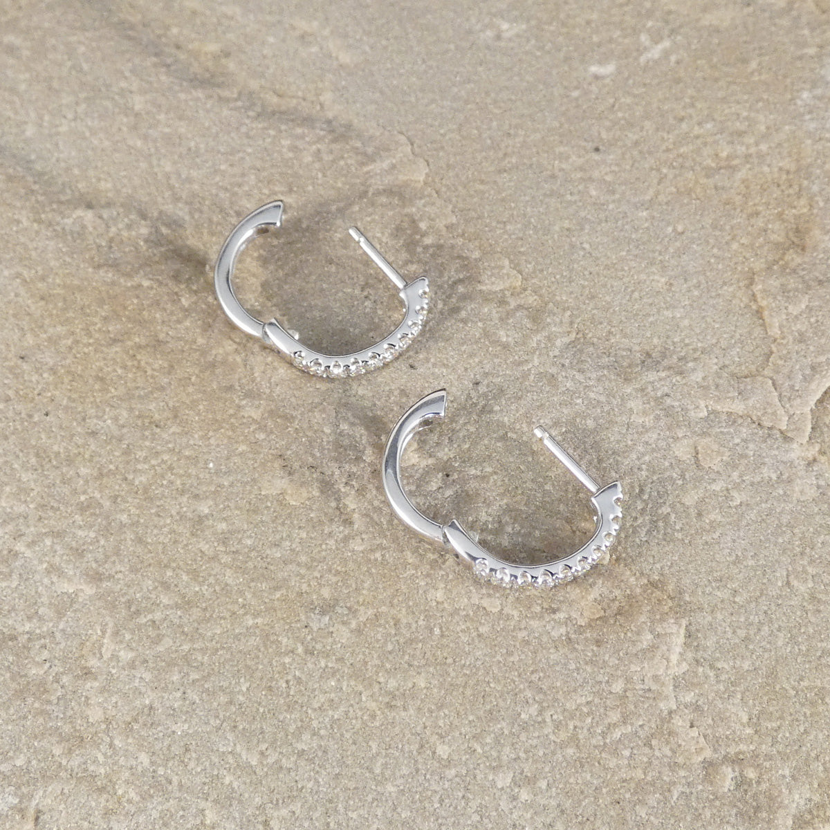 A pair of small 9ct white gold hoop earrings with a row of clear stones along the front, displayed on a textured beige stone surface.