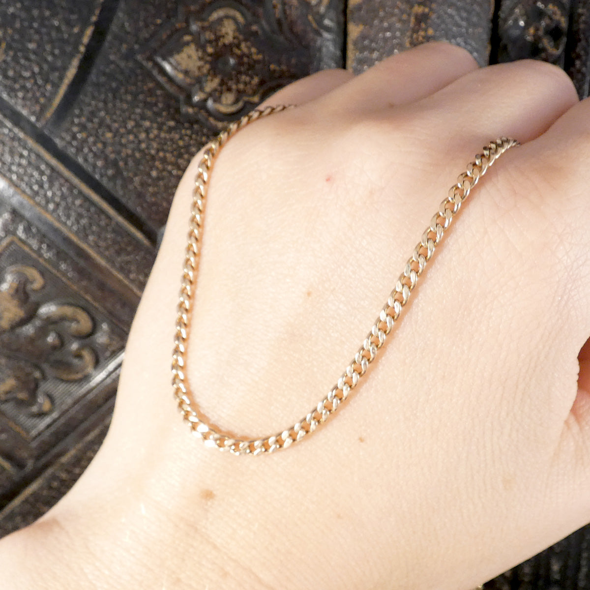 A close-up of a hand wearing a gold chain necklace, resting against a dark textured metal background.