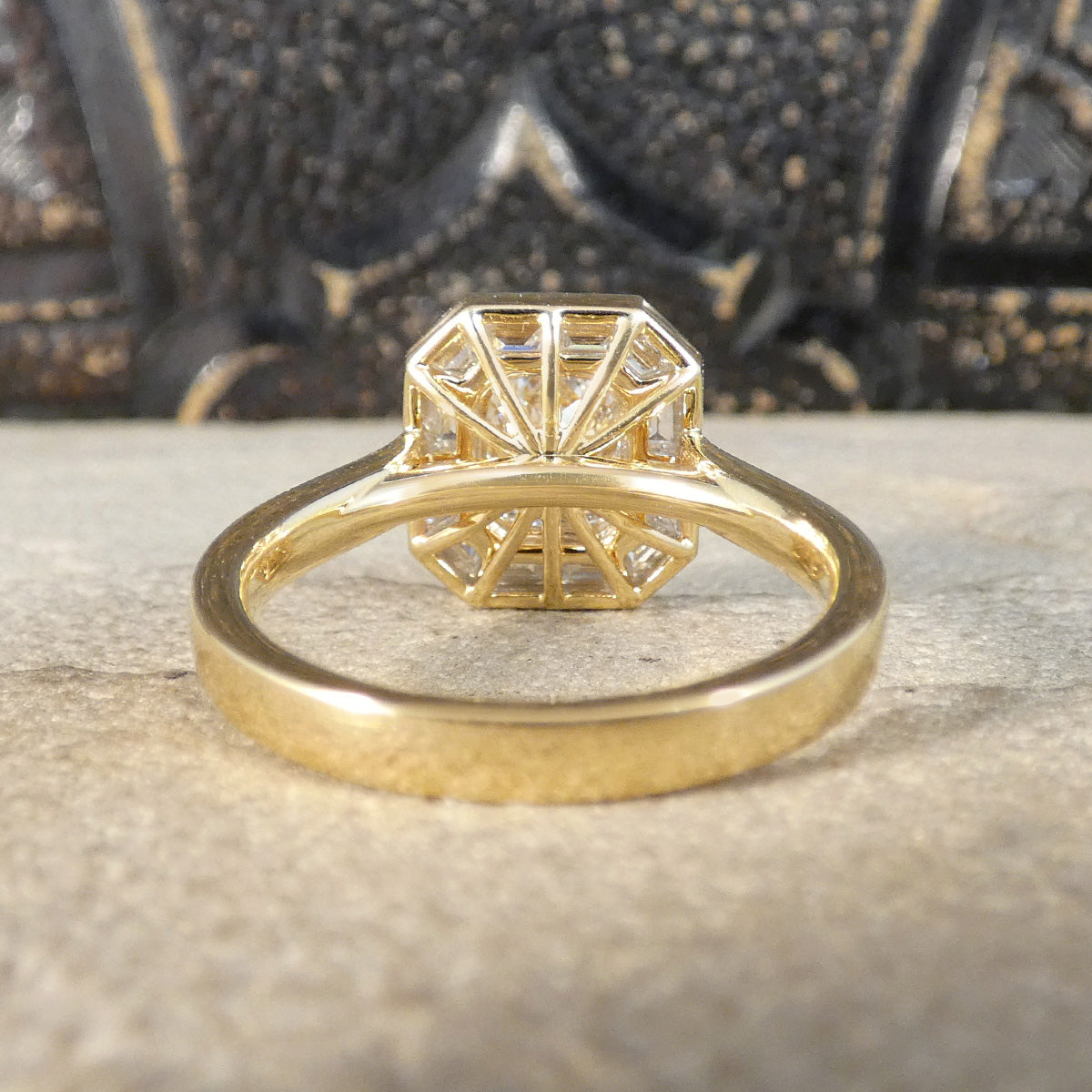 A yellow gold ring with a geometric, fan-like diamond cluster setting, resting on a textured stone surface with a dark, patterned background.