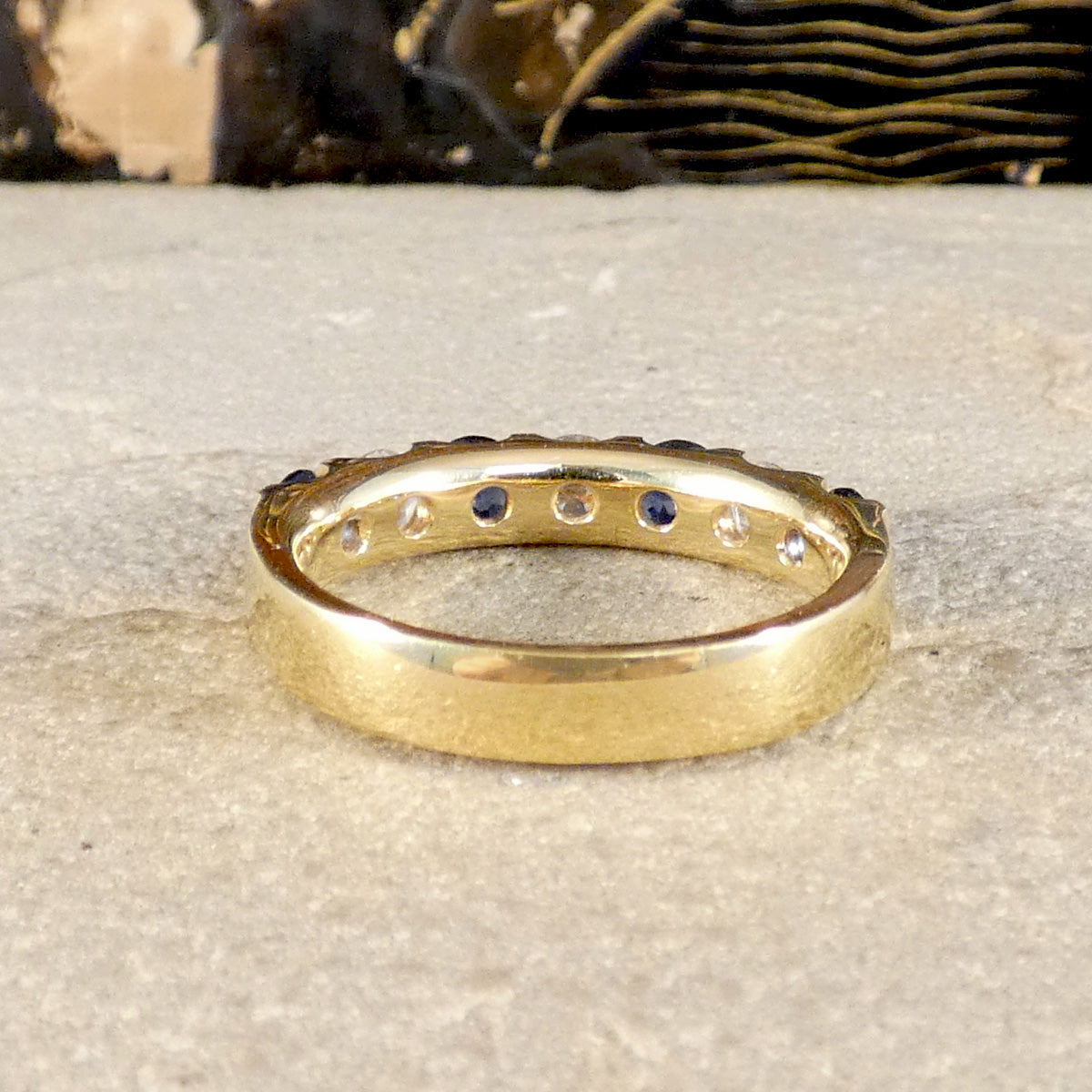 A yellow gold eternity ring with small round clear stones and a row of dark blue sapphires, resting on a textured gray stone surface.