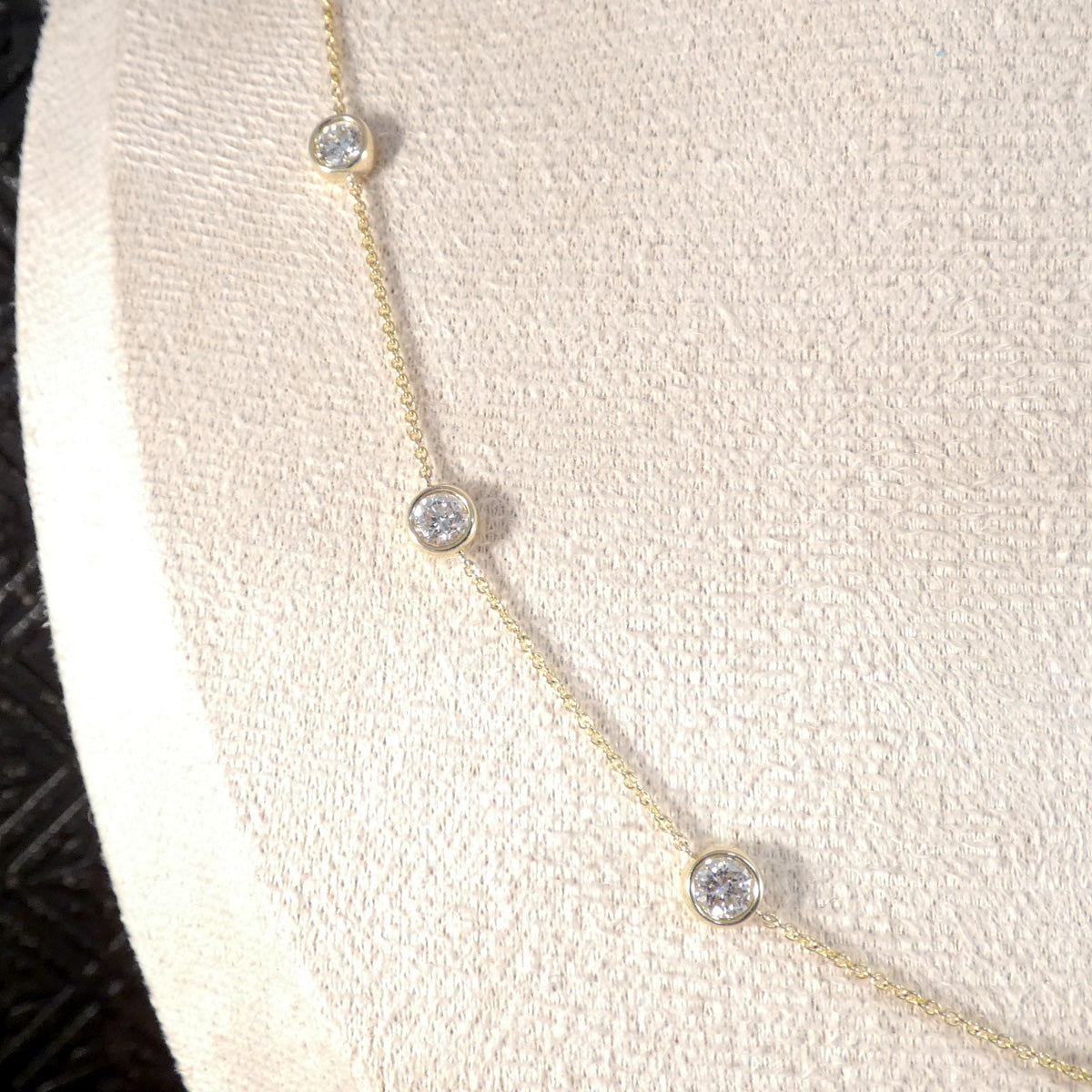 A delicate gold chain necklace with three small round bezel-set clear diamond stones, displayed on a cream-colored textured jewelry bust.