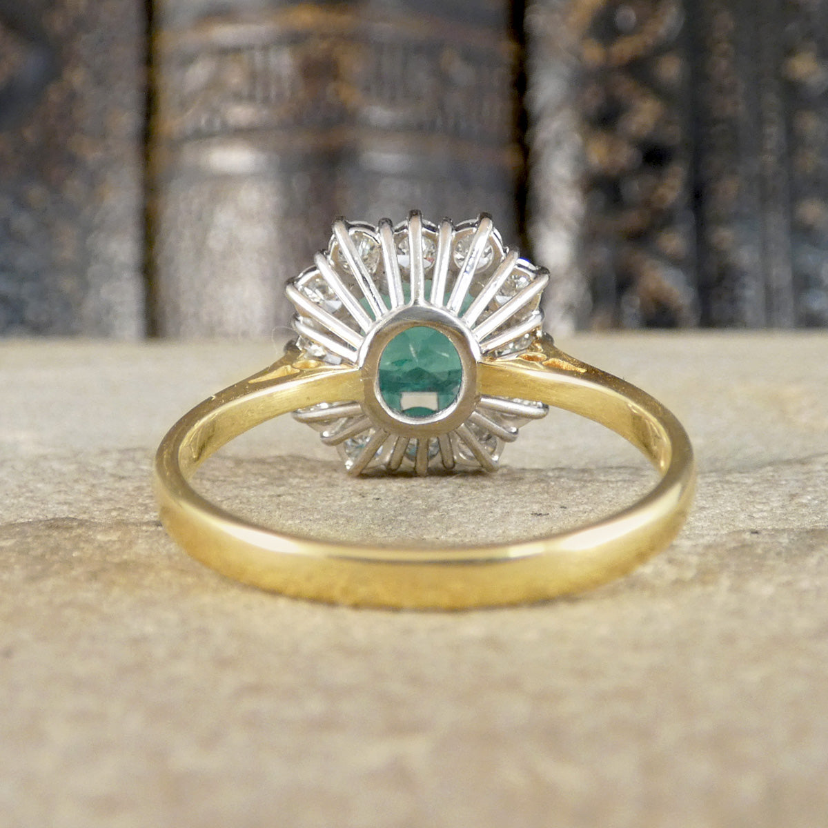 A vintage-style ring with a yellow gold band and a silver-toned sunburst setting featuring a central green emerald, resting on a textured stone surface with blurred books in the background.