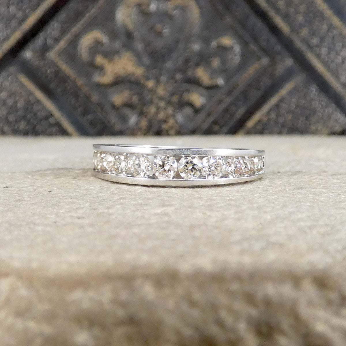 A silver band ring set with multiple round clear diamonds, resting on a textured beige stone surface with a dark decorative background.