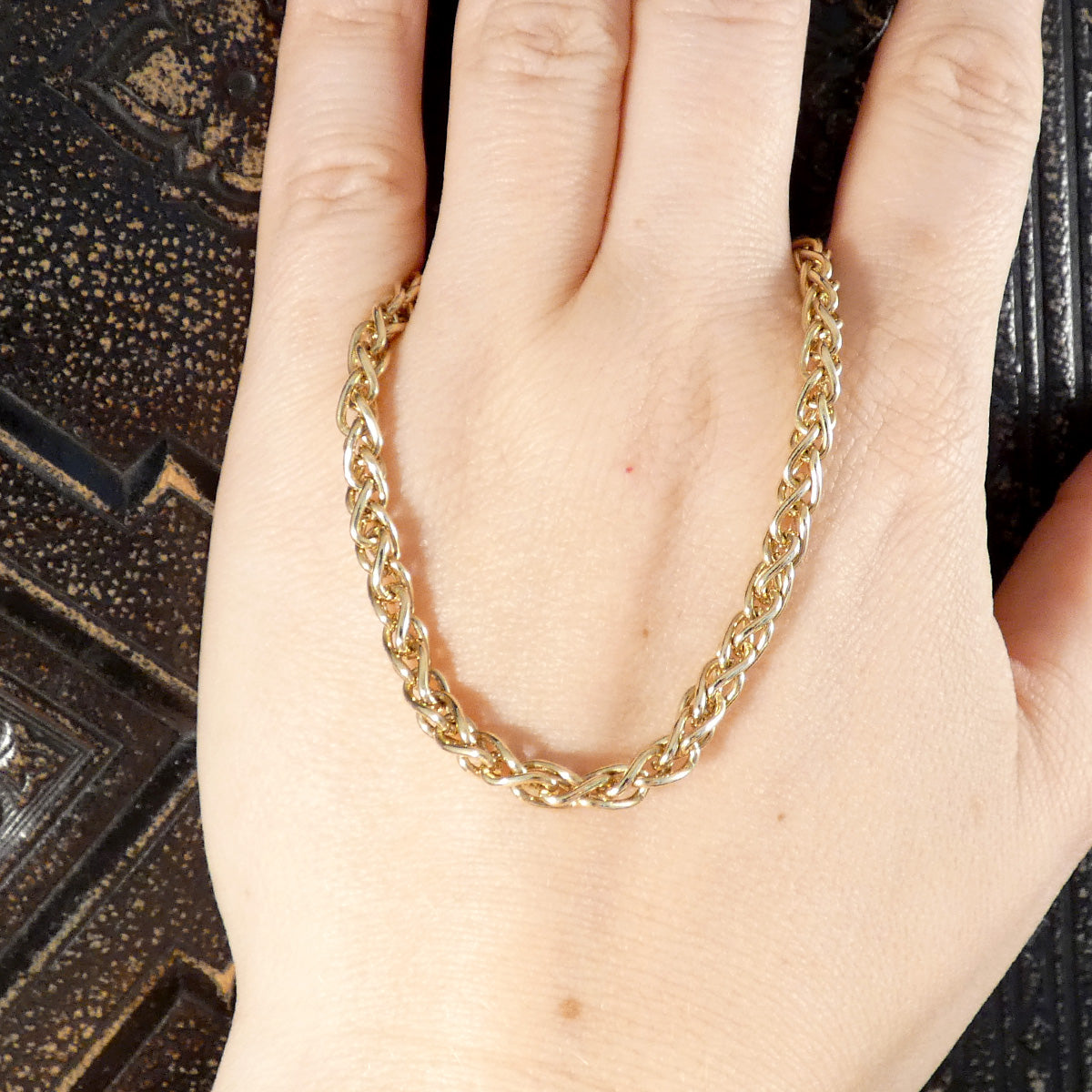 A close-up of a gold Cuban link chain bracelet draped across a person's hand, set against a dark textured background.