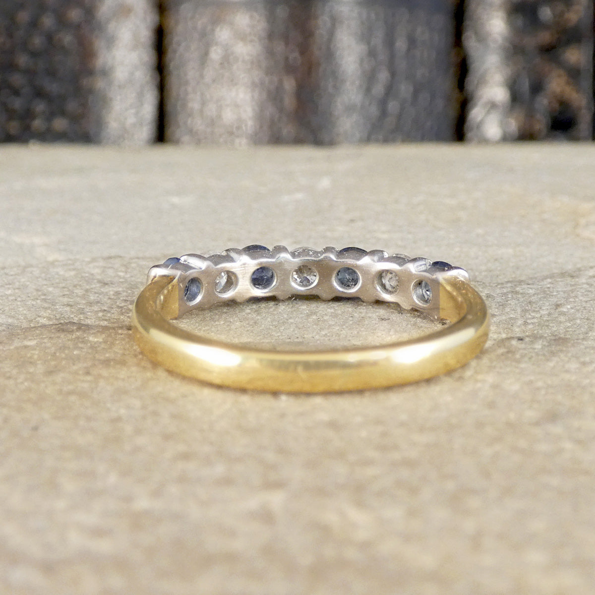 A yellow gold eternity ring with a row of small round-cut blue gemstones, photographed on a textured beige stone surface with a blurred dark background.