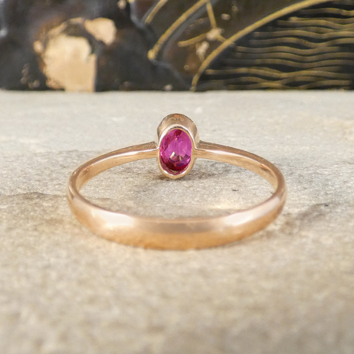 A rose gold ring with a single oval pink gemstone, resting on a textured light gray stone surface with a blurred dark background.