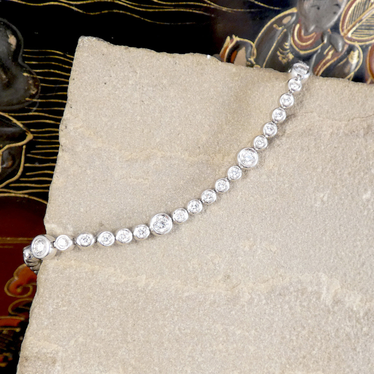 A diamond tennis bracelet with bezel set diamonds of different sizes in white metal on a stone.