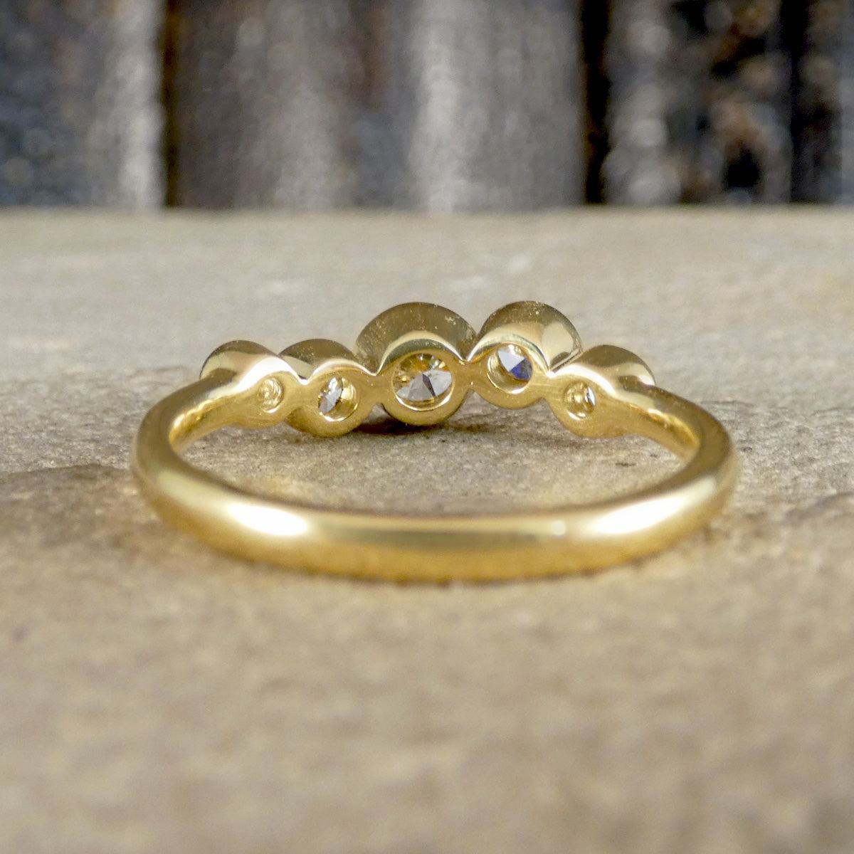 A gold ring with a wavy, scalloped top featuring three round clear gemstones, resting on a textured beige stone surface.