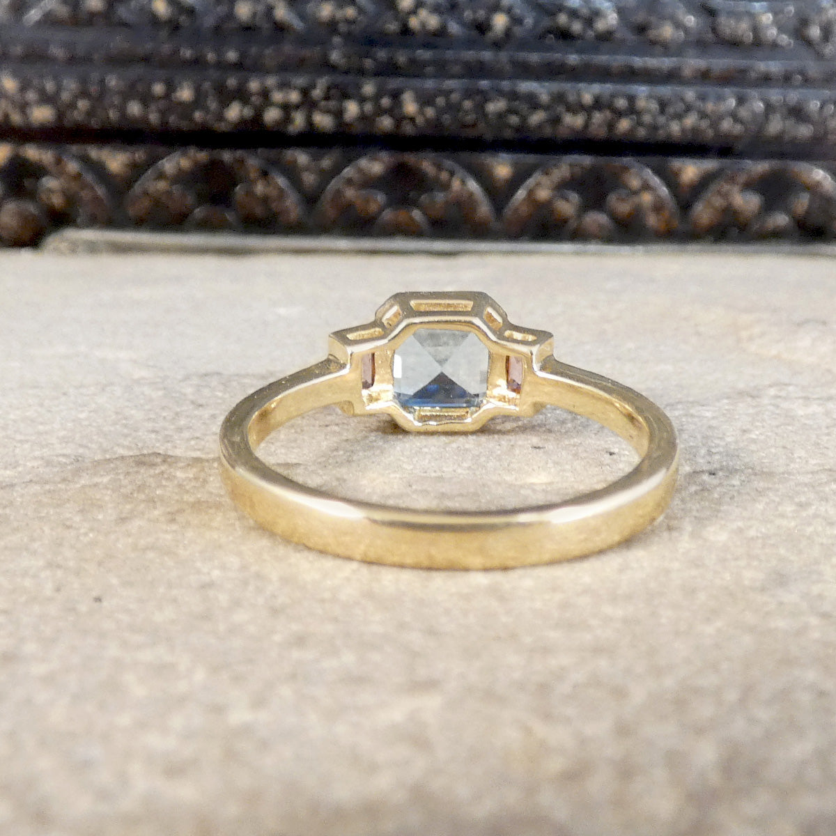 A gold ring with a geometric octagonal bezel holding a clear faceted gemstone, resting on a textured beige stone surface with a dark, ornate background.