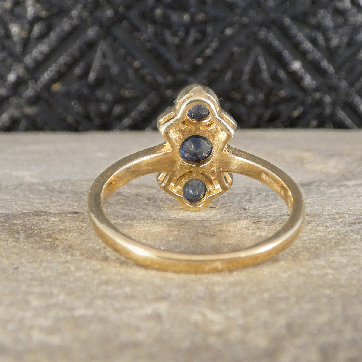 A vintage-style gold ring with a decorative top featuring three dark blue gemstones, displayed on a textured stone surface with a dark patterned background.