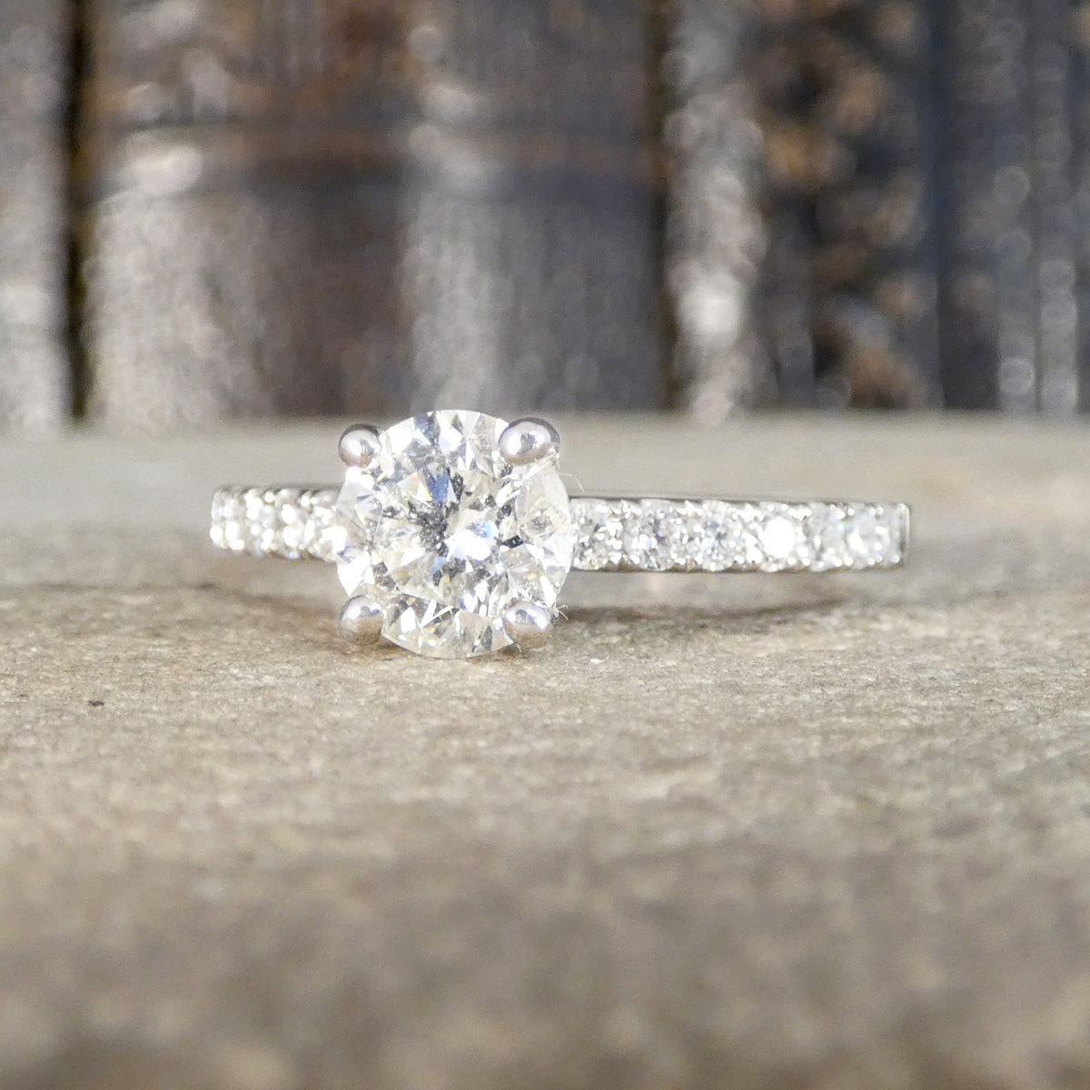 A silver-toned diamond engagement ring with a round-cut center stone and a pavé band, resting on a textured stone surface with a blurred background.
