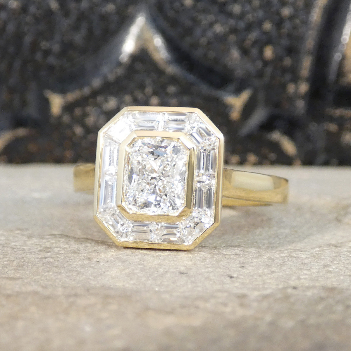 A yellow gold ring featuring an octagonal bezel setting with a central diamond surrounded by baguette and round diamonds, resting on a textured stone surface.