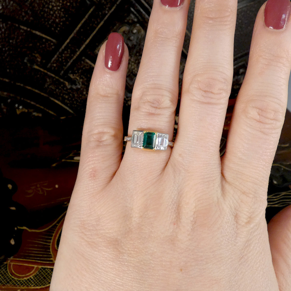 An Emerald and Diamond three stone ring in 18ct white and yellow gold displayed on the ring finger on the hand.