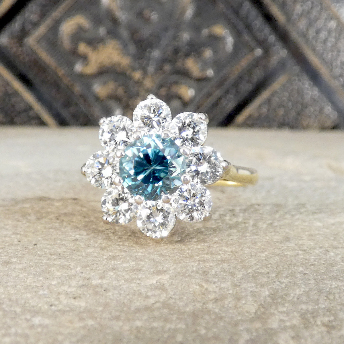 A floral cluster ring featuring a central round-cut light blue gemstone surrounded by a halo of clear round stones, set on a yellow gold band and displayed on a textured stone surface.