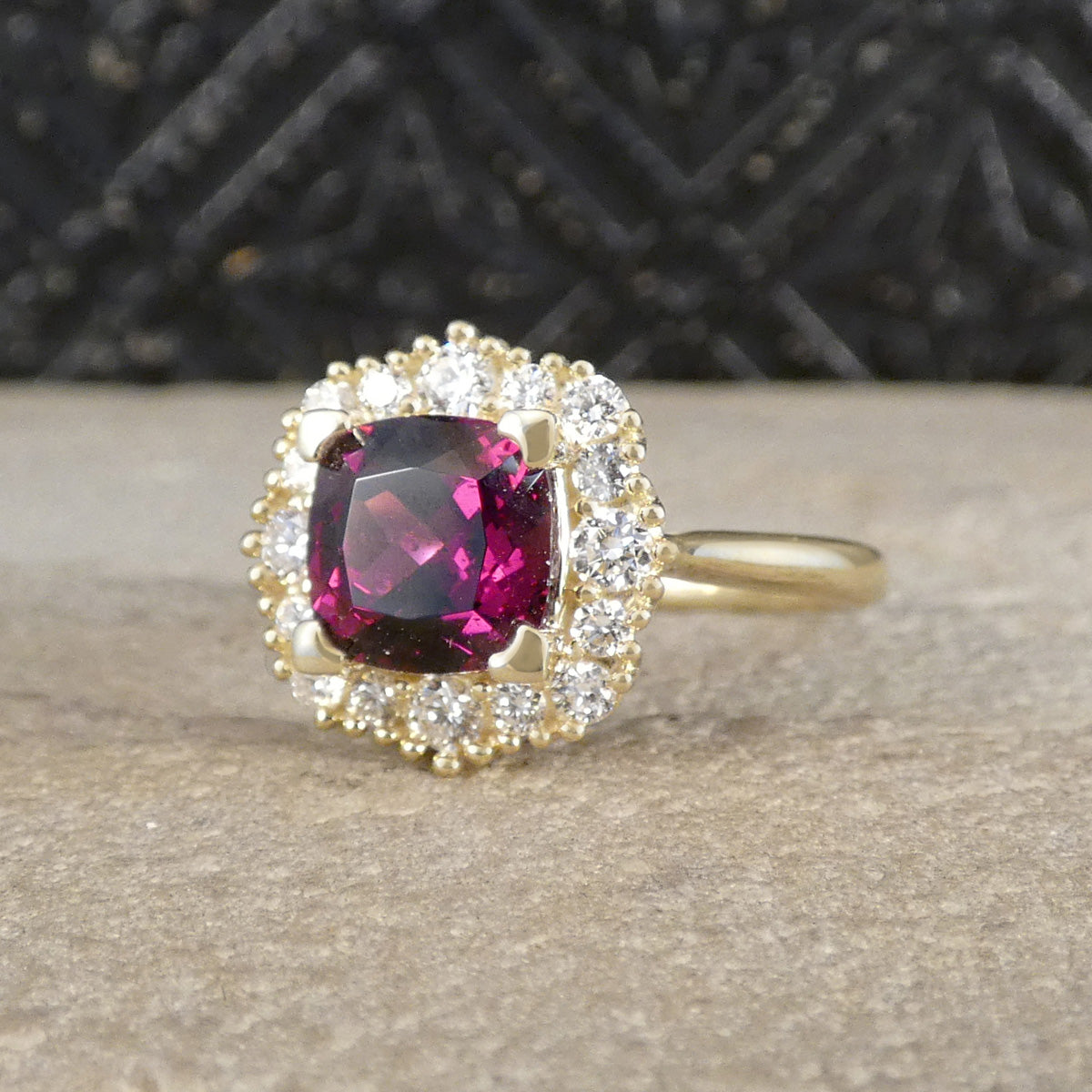 A yellow gold ring featuring a cushion-cut dark pinkish-red center stone surrounded by a halo of small clear stones, displayed on a textured beige surface with a dark patterned background.