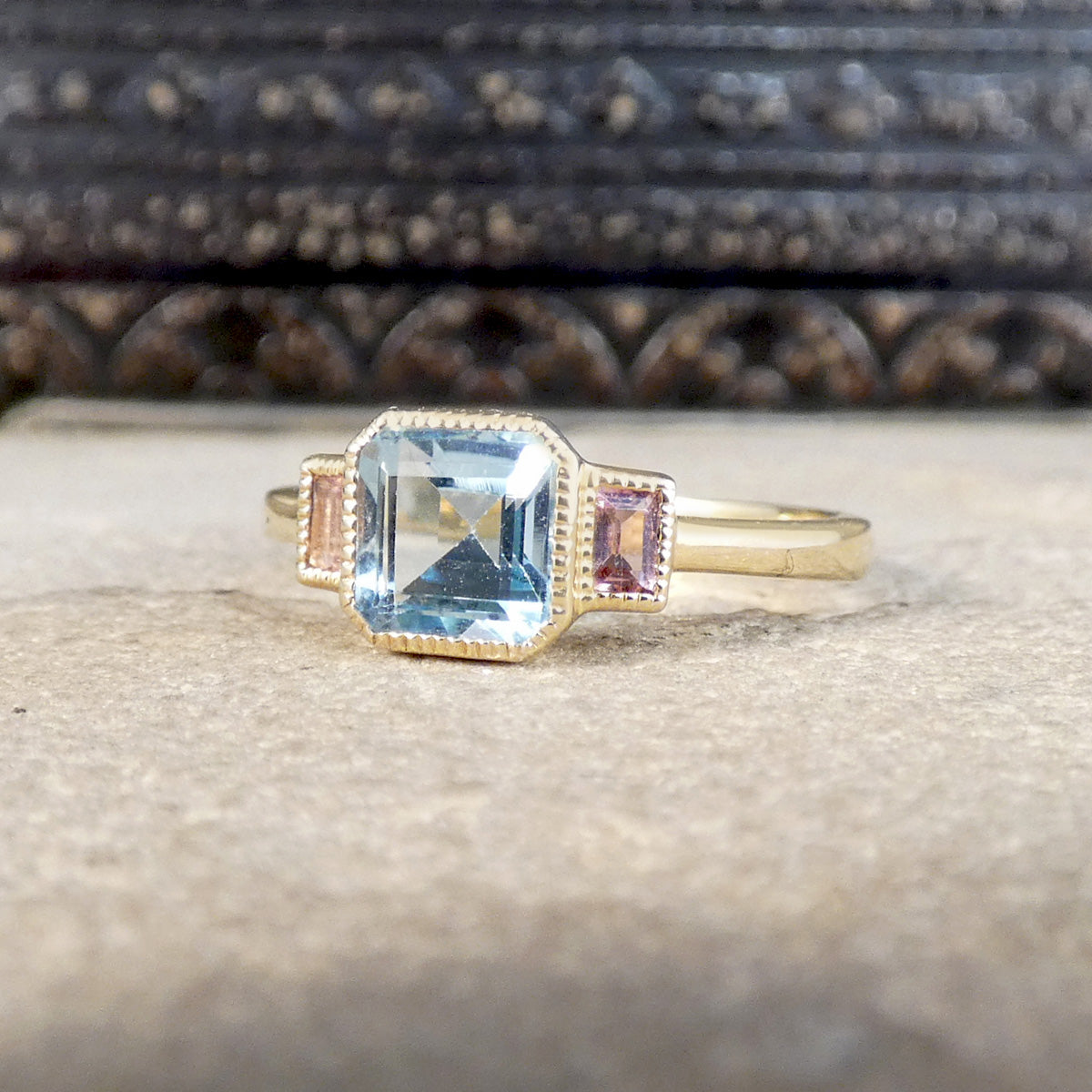 A gold ring featuring a large emerald-cut light blue topaz center stone, accented by two small pinkish-orange baguette-cut stones on either side, displayed on a textured stone surface.