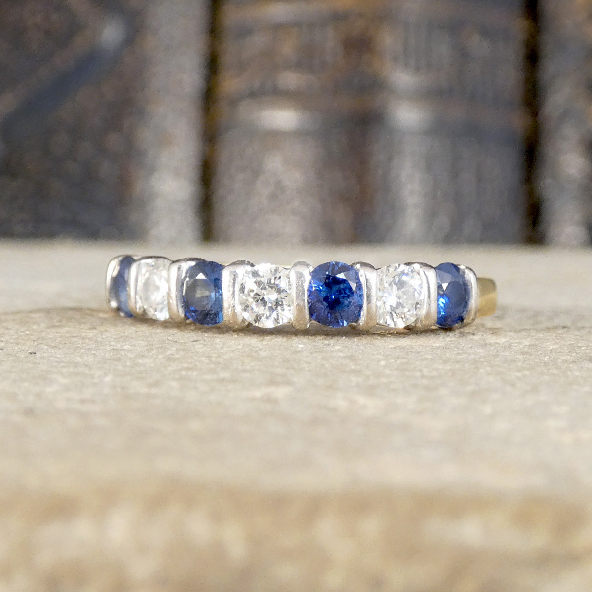 A vintage-style eternity ring featuring alternating round blue sapphires and clear round diamonds in a silver-toned setting, resting on a textured beige stone surface with a blurred dark background.
