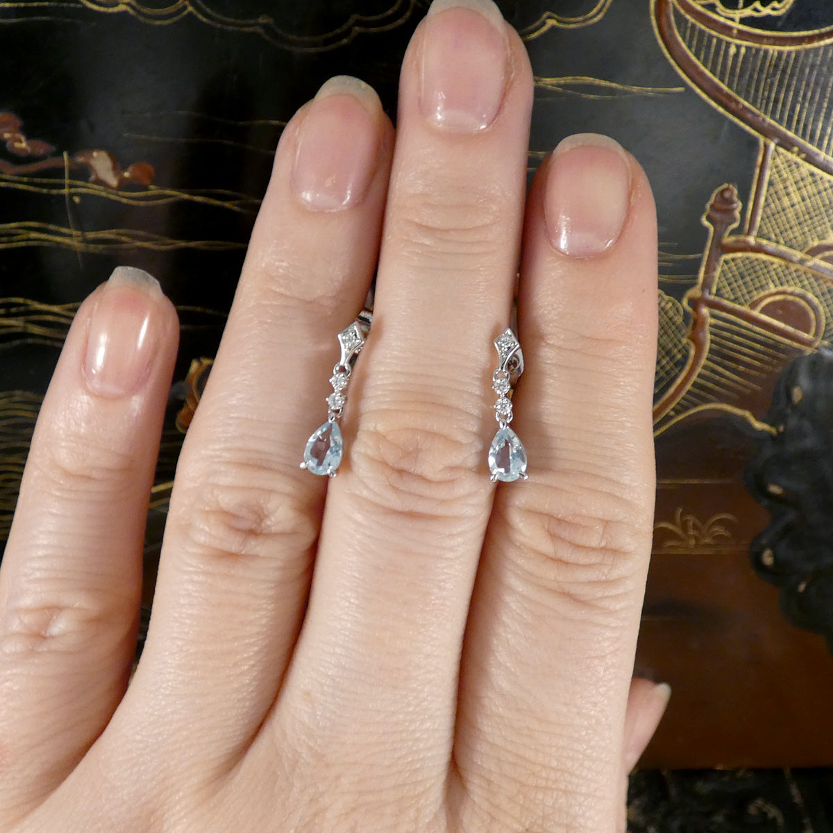 Aquamarine and Diamond teardrop earrings on a hand with an ornate background