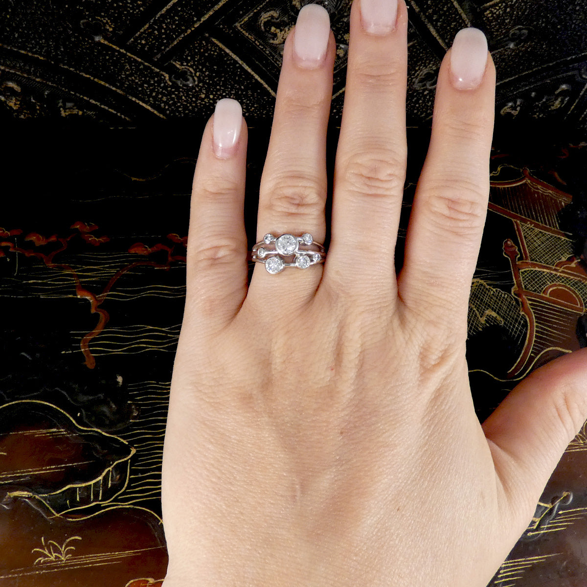 A silver-toned three-stone ring with clear gemstones worn on a hand with pale pink manicured nails, set against a dark patterned background.