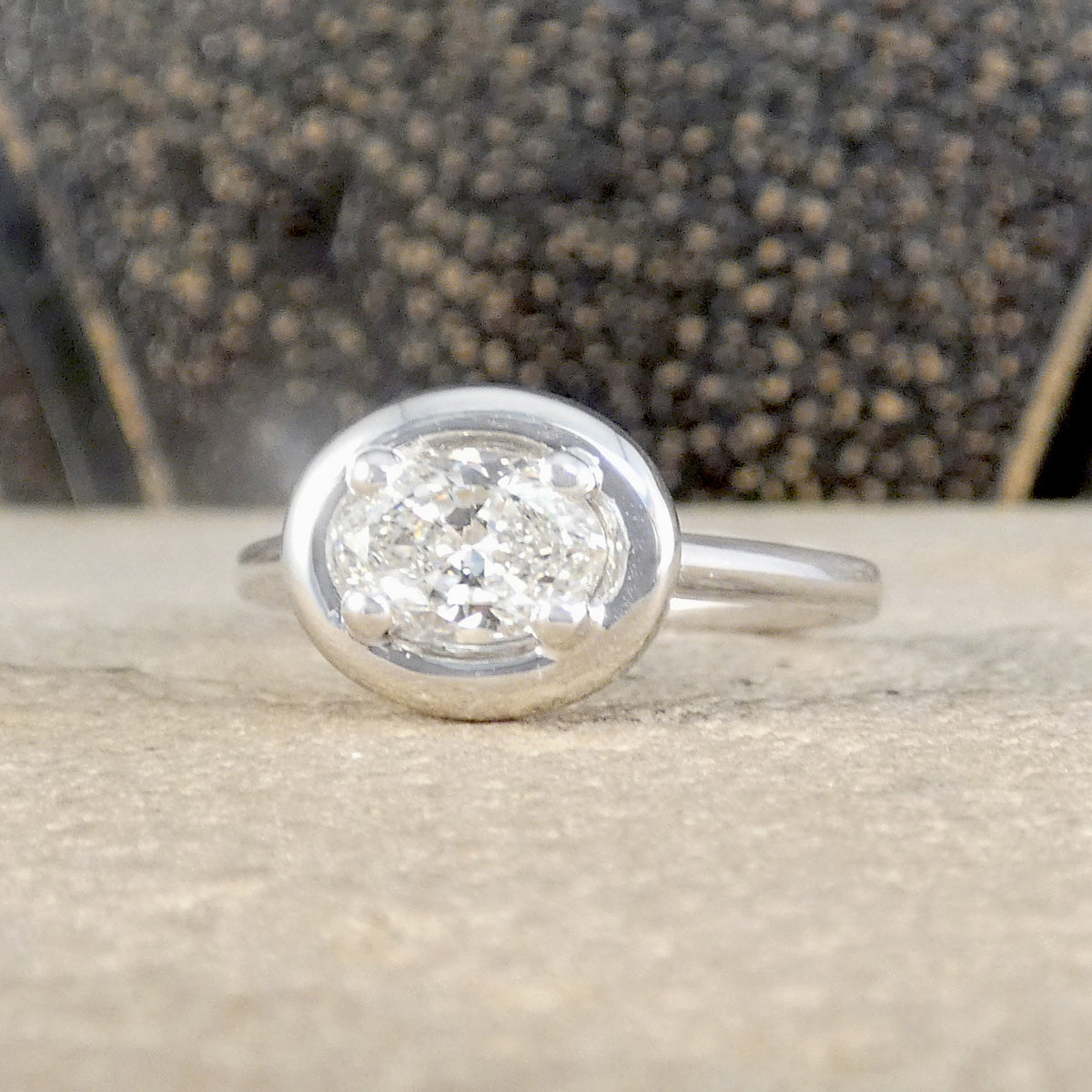 A silver ring with an oval-cut clear gemstone in a prong setting, resting on a textured beige stone surface with a dark, out-of-focus background.