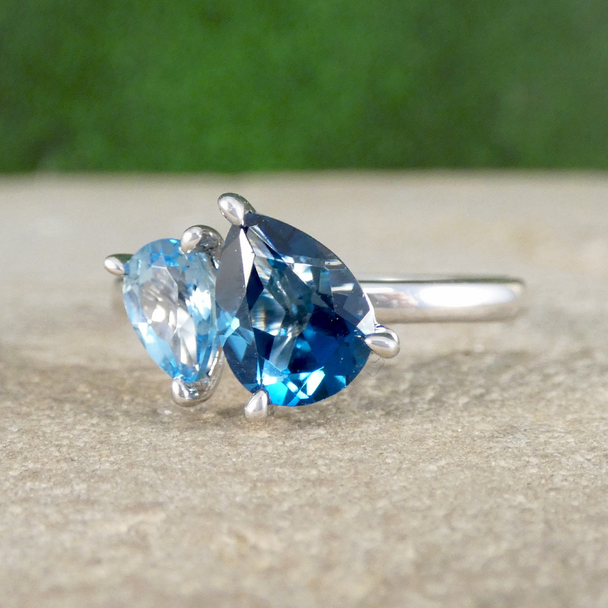 White Gold ring with pear-shaped blue gemstones on a textured surface with a blurred green background