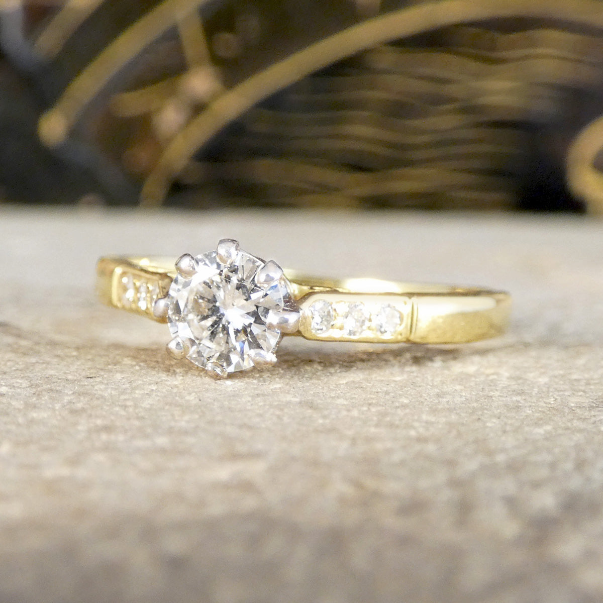 A yellow gold solitaire engagement ring with a round-cut diamond center stone and small accent diamonds on the band, resting on a textured stone surface.
