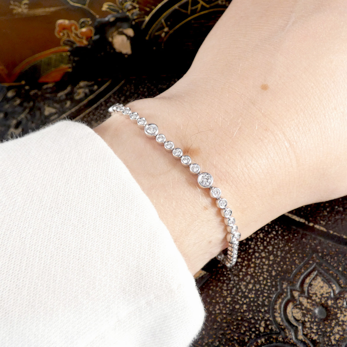 A diamond tennis bracelet with bezel set diamonds of different sizes in white metal shown on the wrist