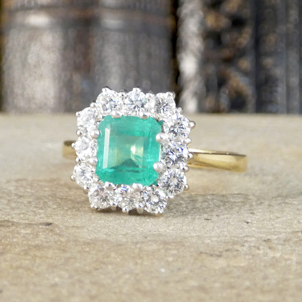 A yellow gold ring featuring a square-cut green emerald center stone surrounded by a halo of clear cubic zirconia, resting on a textured stone surface with a blurred background.
