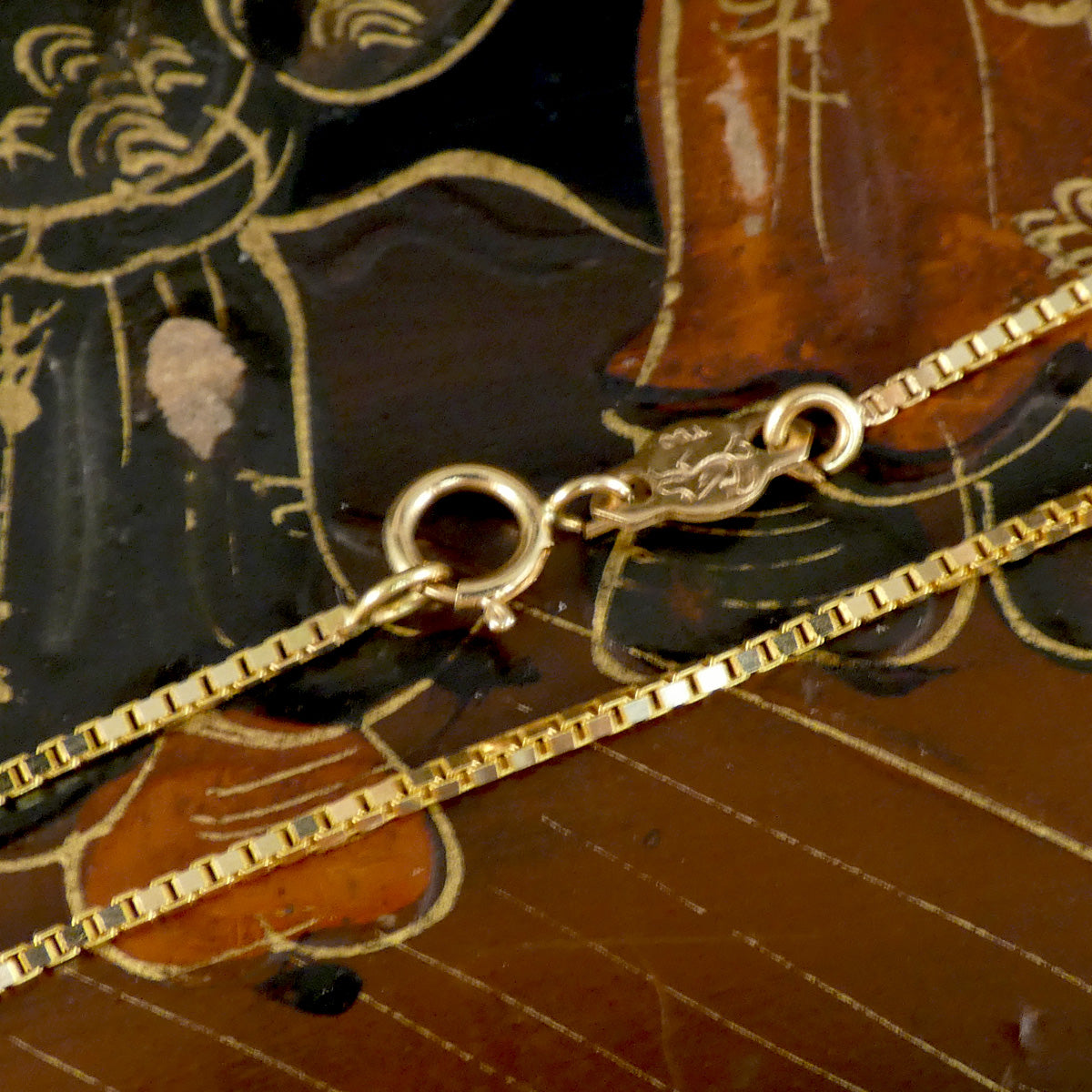 A close-up of a gold box chain necklace with a lobster clasp, laid diagonally across a dark brown and black painted wooden surface.
