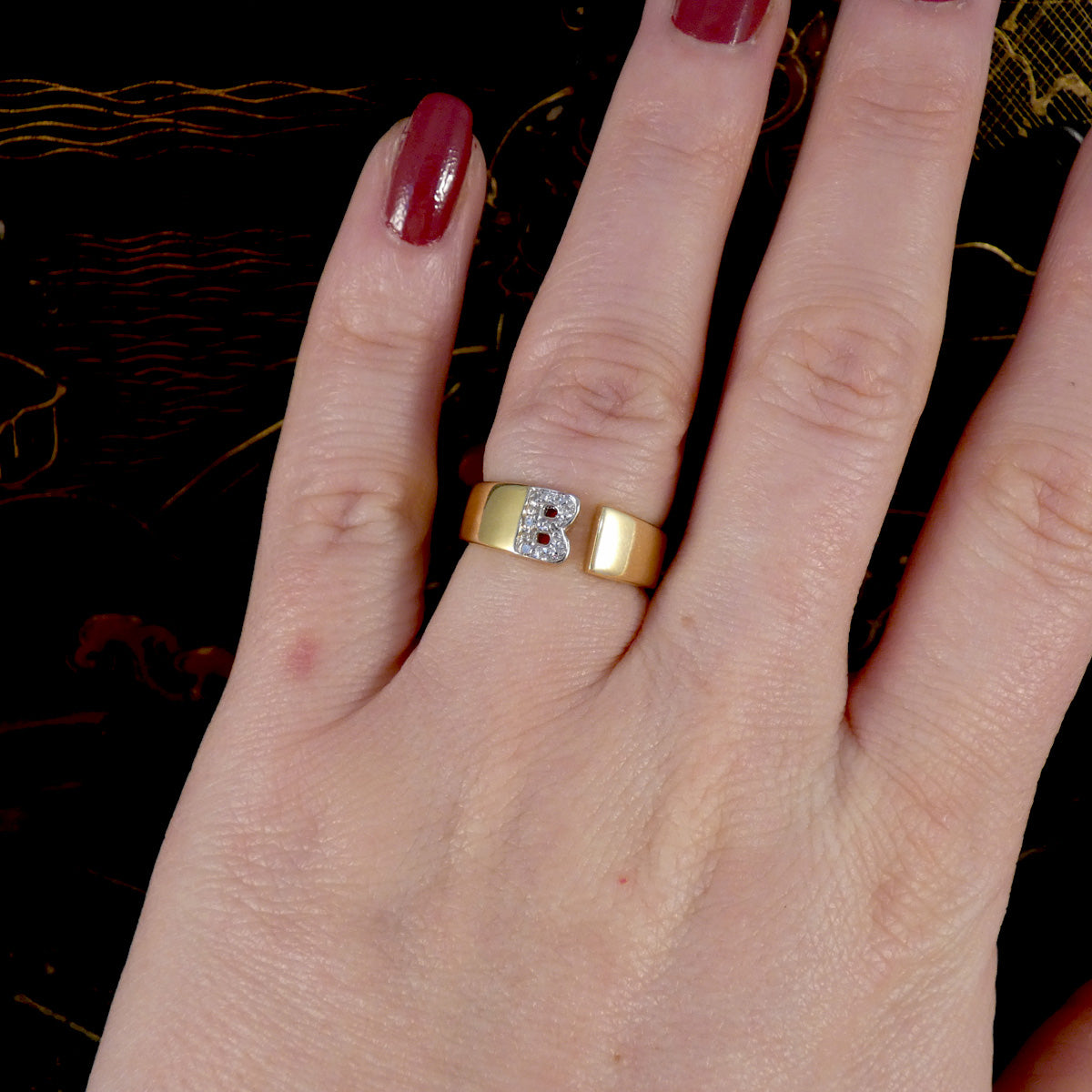 Hand wearing a gold ring with a 'B' on a dark background