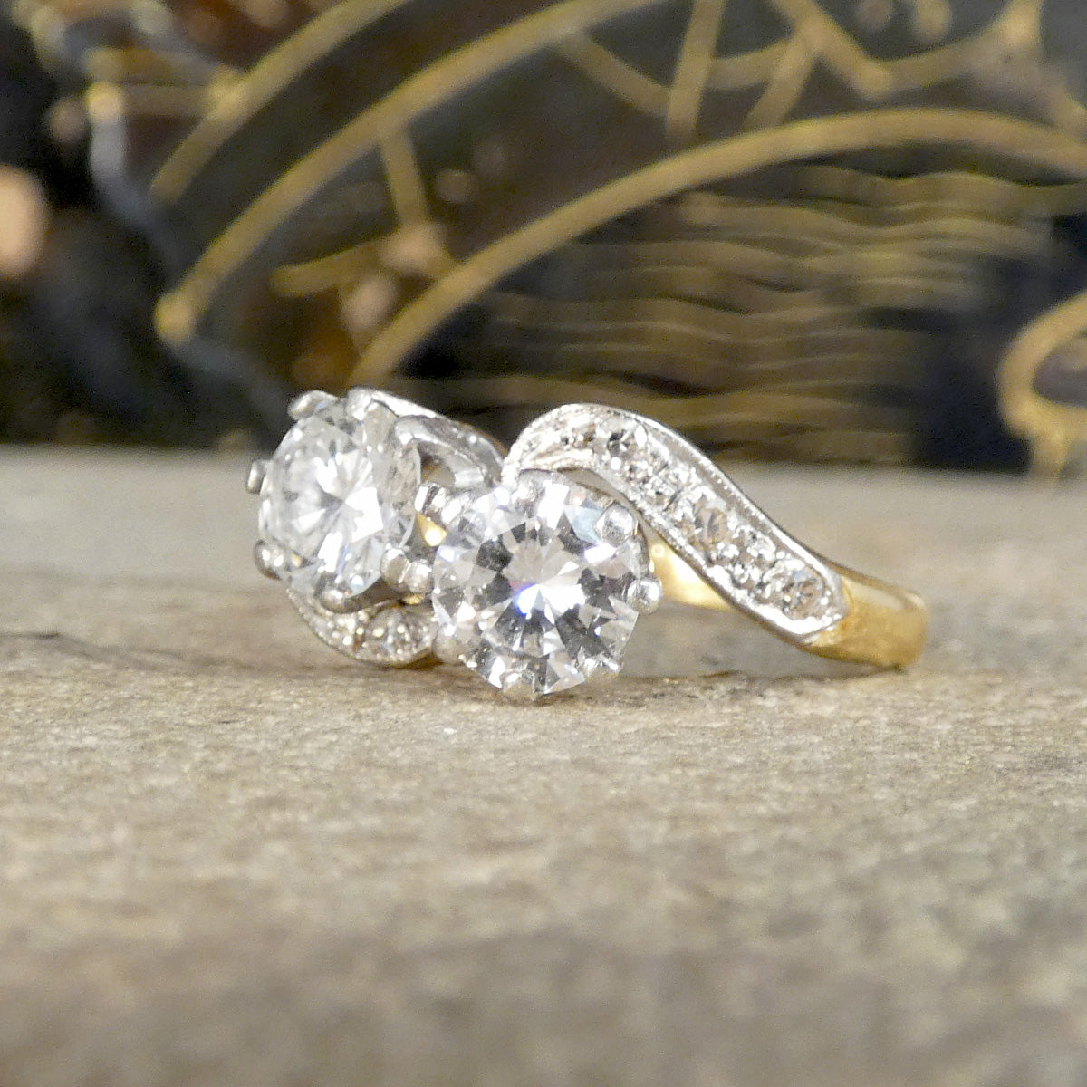 A three-stone diamond engagement ring with a silver-toned pavé band and a yellow gold section, resting on a textured stone surface.