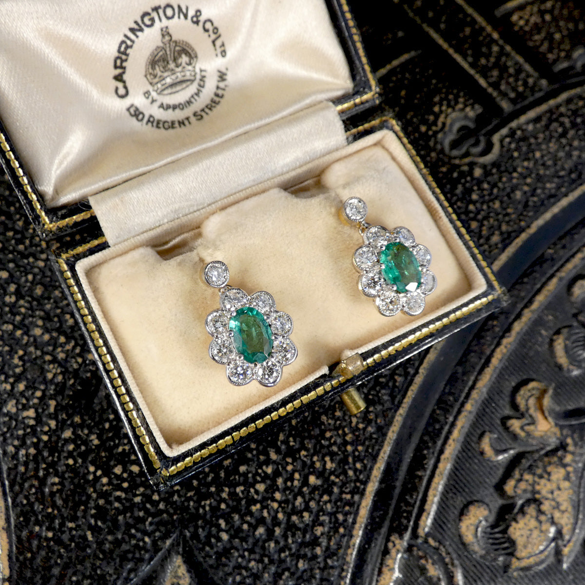 A pair of Carrington & Cels earrings with oval green emerald-cut gemstones and surrounding clear stones, presented in an open vintage-style jewelry box with cream velvet lining.