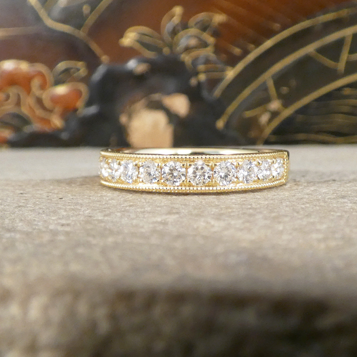 A yellow gold eternity ring set with round brilliant-cut diamonds, resting on a textured gray stone with a blurred decorative background.