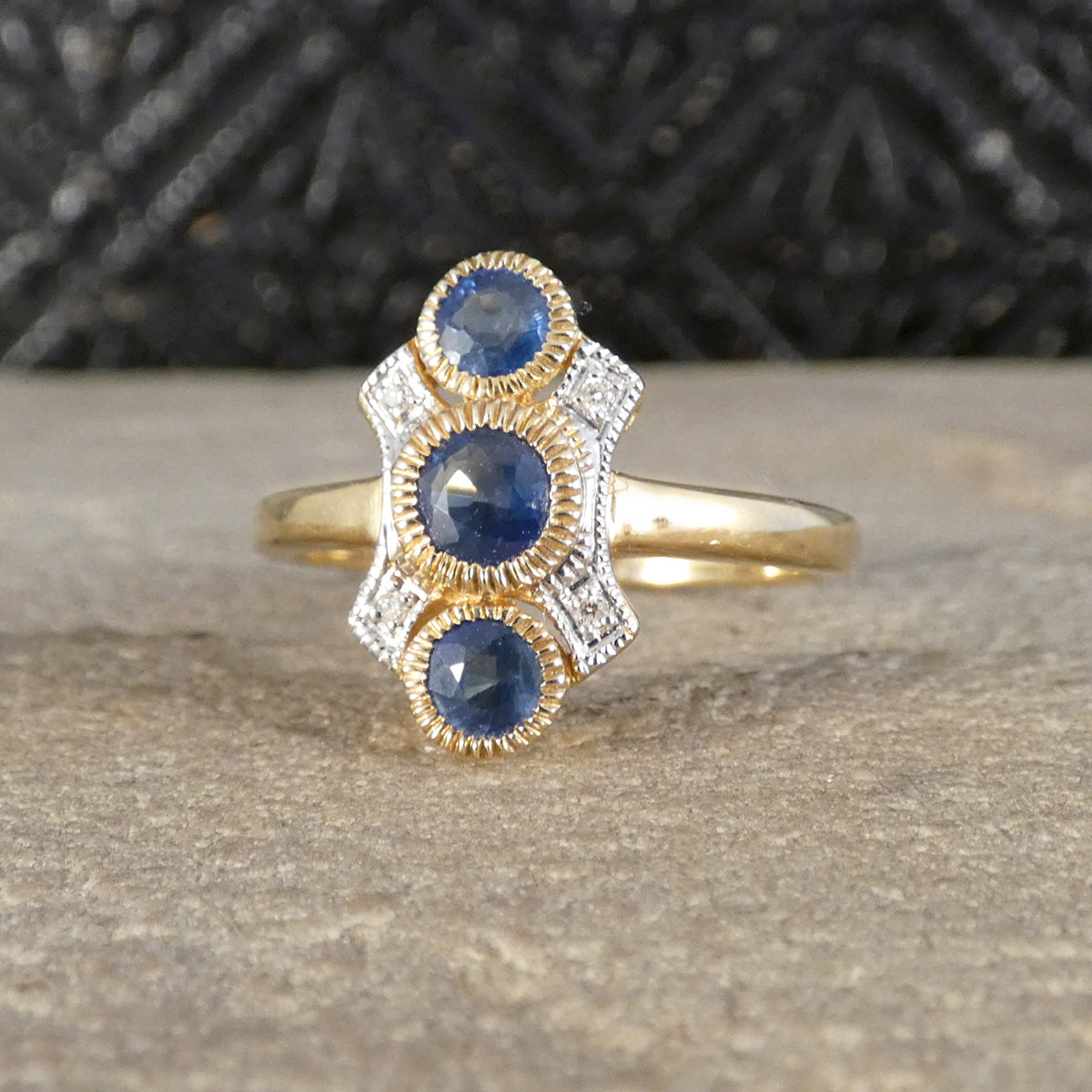 A gold and white gold ring featuring three round blue sapphires in a decorative Art Deco style setting with small clear accent stones, resting on a textured gray stone surface.