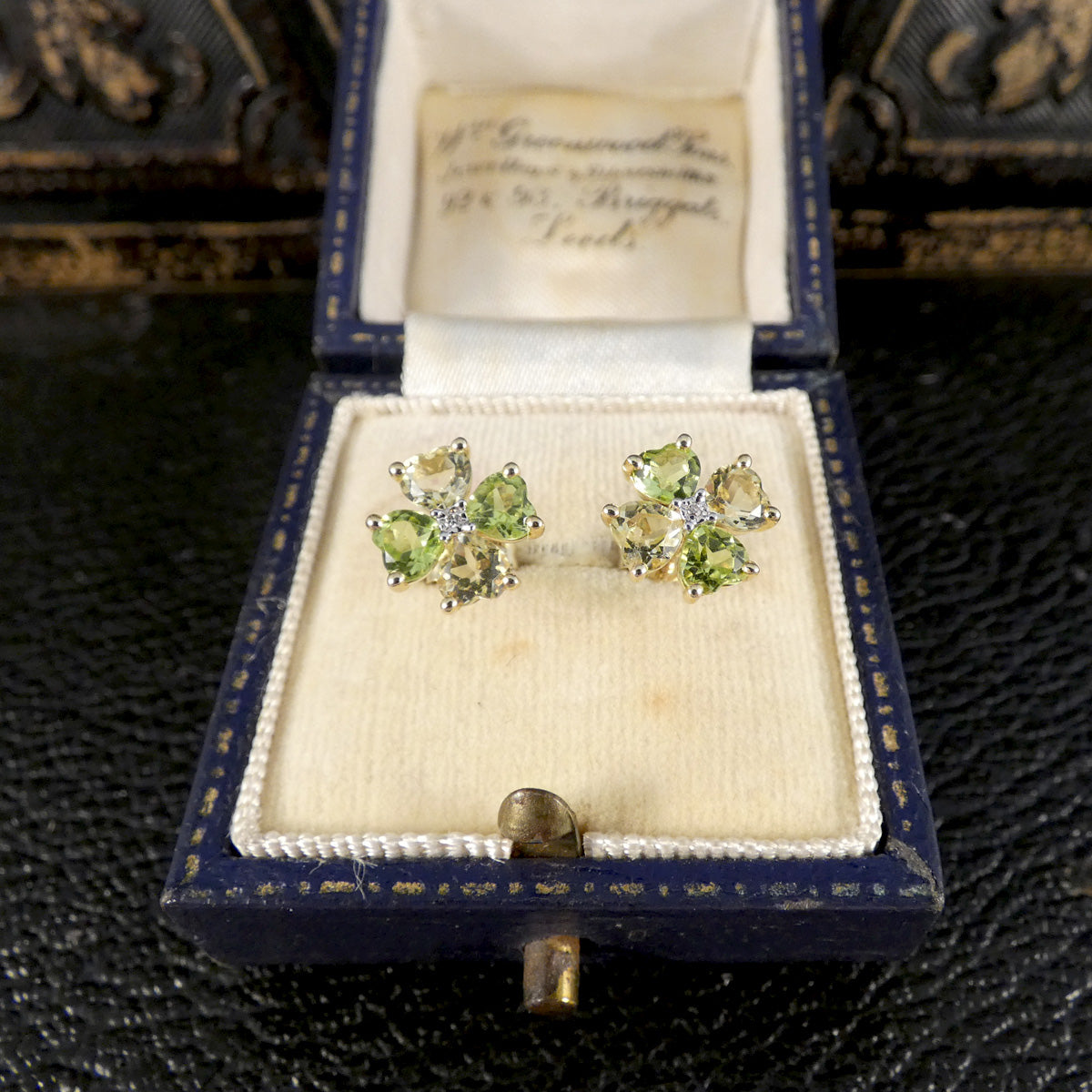 A pair of yellow gold stud earrings with four green and yellow gemstones arranged in a flower shape, in jewellery box.