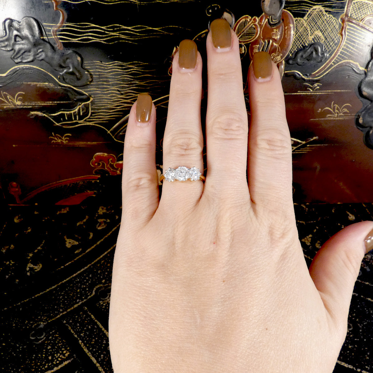 A close-up of a hand wearing a silver-toned ring with a curved row of clear gemstones, set against a dark lacquered wooden background with gold and red patterns.