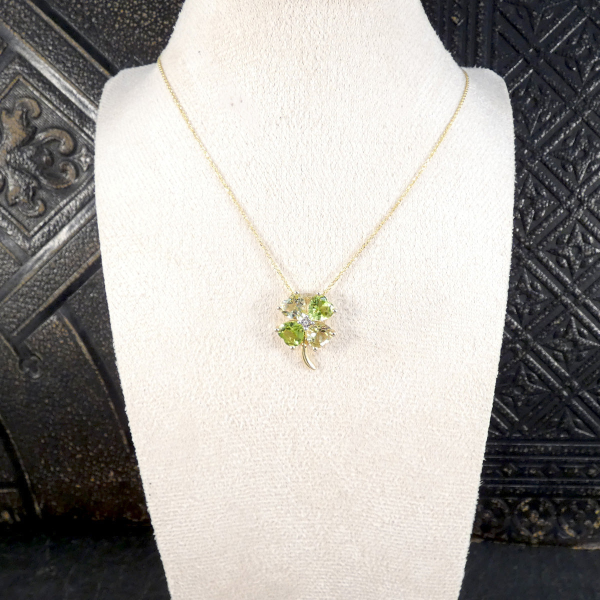 A gold necklace with a four-leaf clover pendant featuring green and clear crystals, displayed on a white fabric jewelry bust.