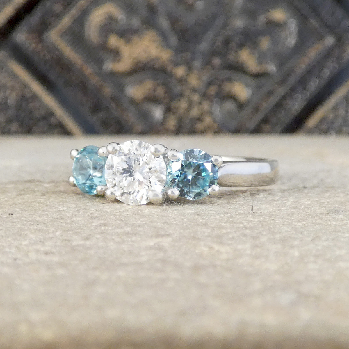 A silver three-stone ring featuring a round brilliant-cut center stone and two round light blue side stones, displayed on a textured stone surface with a dark decorative background.