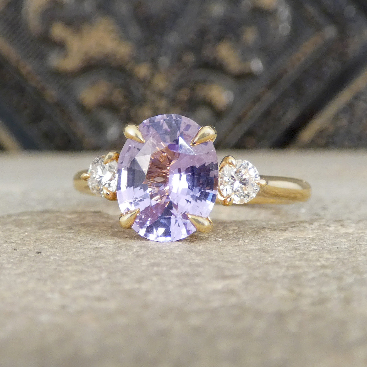 A yellow gold ring featuring an oval-cut light purple sapphire center stone, accented by two round clear diamonds on either side, resting on a textured stone surface with a dark, ornate background.