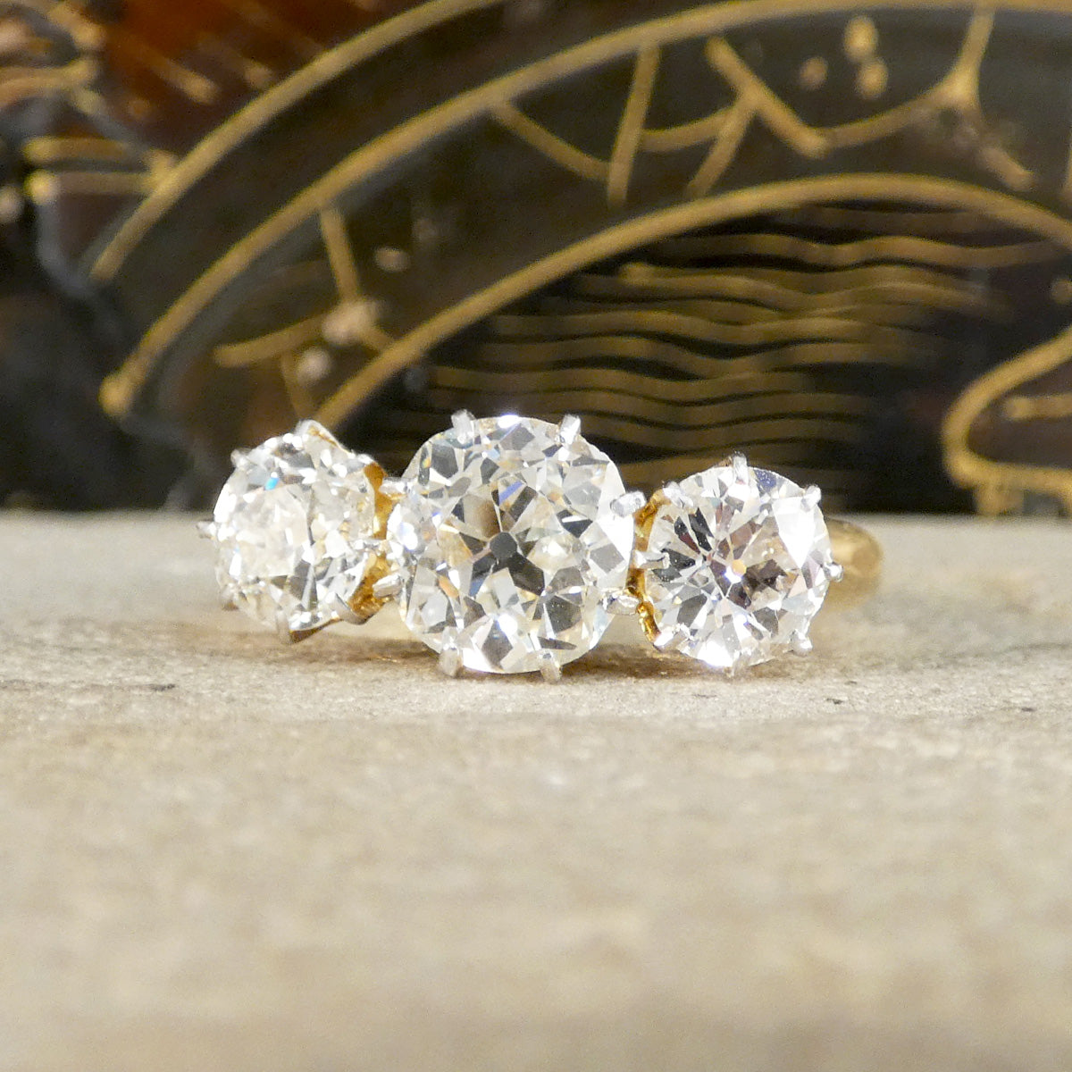 Three-stone diamond ring with gold setting on a textured surface.