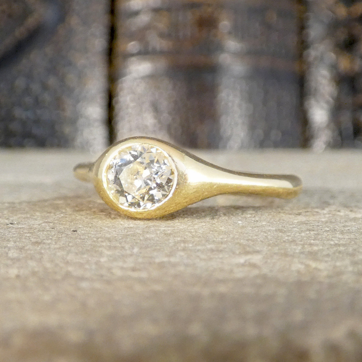 A gold ring with a large round clear gemstone in a bezel setting, resting on a textured stone surface with a blurred dark background.