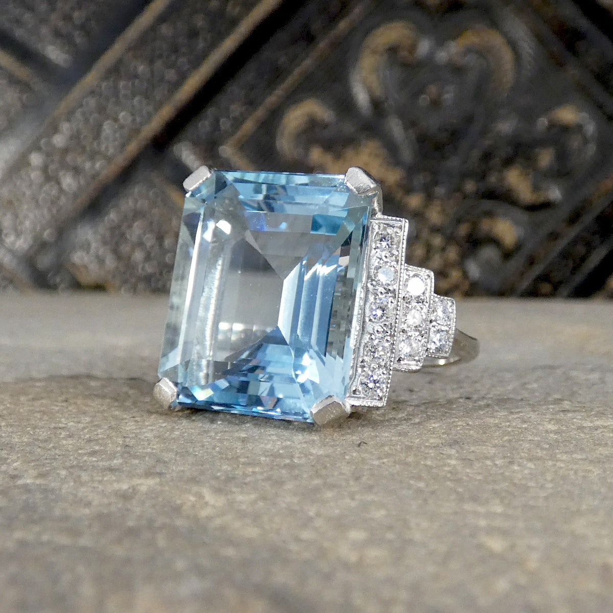 An emerald-cut light blue aquamarine gemstone ring set in a silver-toned metal with pavé-set clear stones, resting on a textured gray stone surface with a dark ornate background.