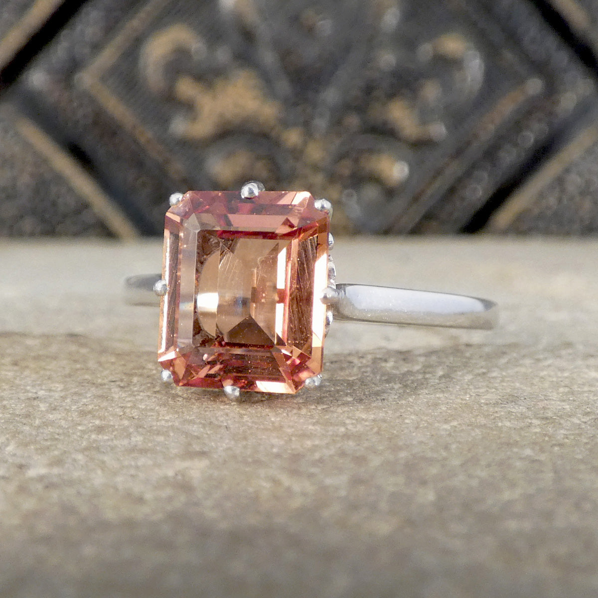 An emerald-cut orange-colored gemstone ring set in a silver band, resting on a textured stone surface with a blurred dark metal background.