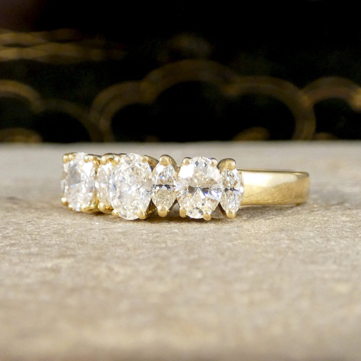 A yellow gold five-stone diamond ring with oval and marquise-cut diamonds, resting on a textured beige surface with a dark, blurred background.