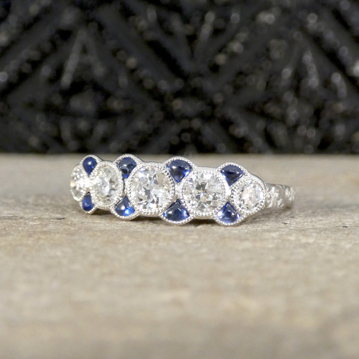 Art Deco-inspired diamond and sapphire ring in platinum, featuring five old-cut diamonds accented by vivid blue triangle-cut sapphires in a milgrain setting, creating a vintage eternity band style.