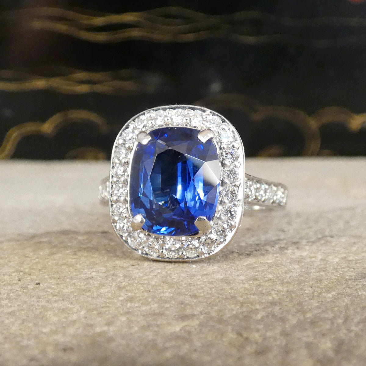 A cushion-cut sapphire halo ring in silver-toned metal, featuring a prominent deep blue sapphire center stone surrounded by a halo of small clear stones, displayed on a textured stone surface.